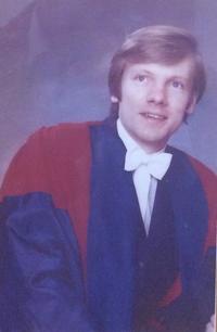 Image of Michael Lockwood, posing for a formal photograph in subfusc, with a red and blue academic gown